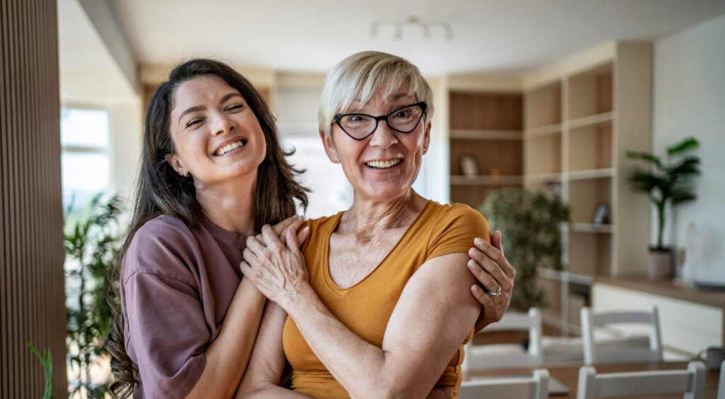 A mother and adult daughter embrace and smile at home, sharing a warm, joyful moment that highlights multi-generational love, connection and everyday family togetherness.