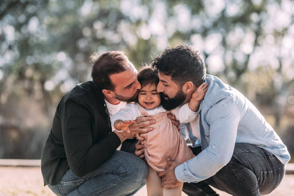 Gay couple kissing their daughter who is smiling amusedly in an urban park.