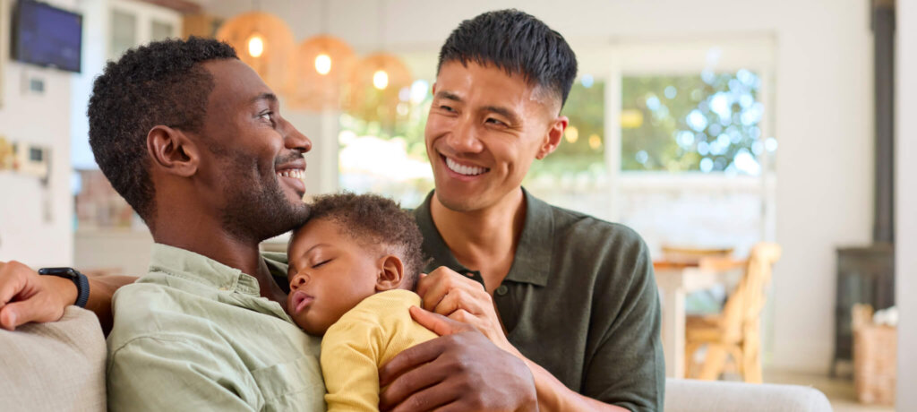 A gay couple cuddles their sleeping baby.